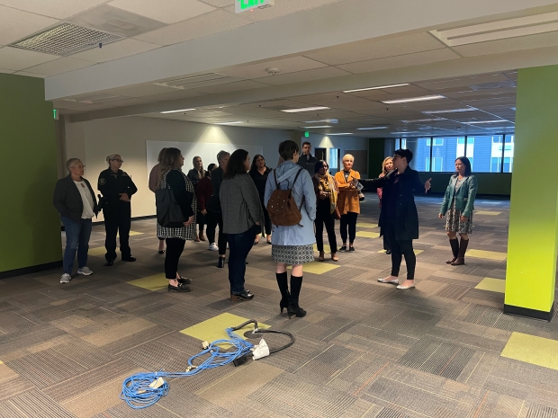 photo of interior of a building with gray patterned carpet flooring, with a group of people looking at an individual gesturing around the building during a tour