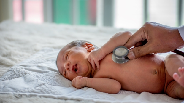 newborn laying on white sheet, with a doctor gently pressing a stethoscope on the newborn's abdomen