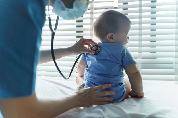 baby sitting on a bed, with a doctor gently pressing a stethoscope on the baby's back