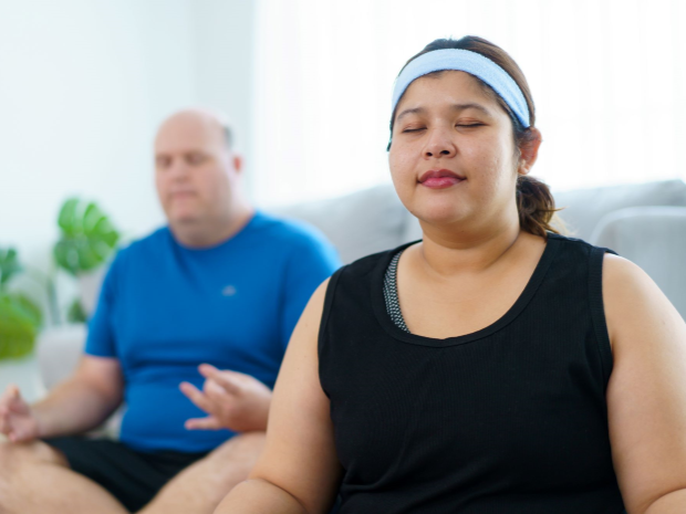 Two people pictured, one in foreground and one in background, both meditating with their eyes closed